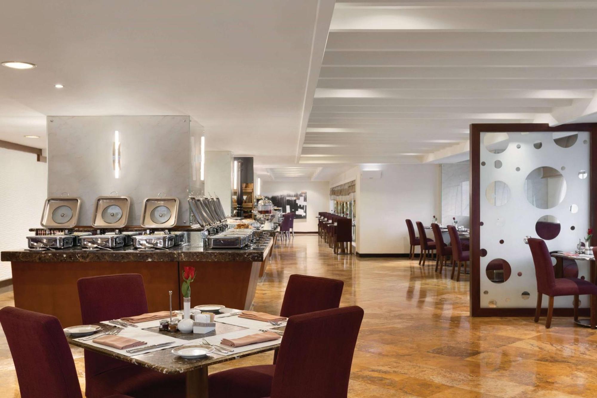 Grand Regency Doha, Trademark Collection By Wyndham Exterior photo The photo shows a modern restaurant interior. There are several tables with maroon chairs arranged throughout the space. One table is set with a small vase containing a flower and dining utensils. Along one side, there is a buffet area with a sleek c