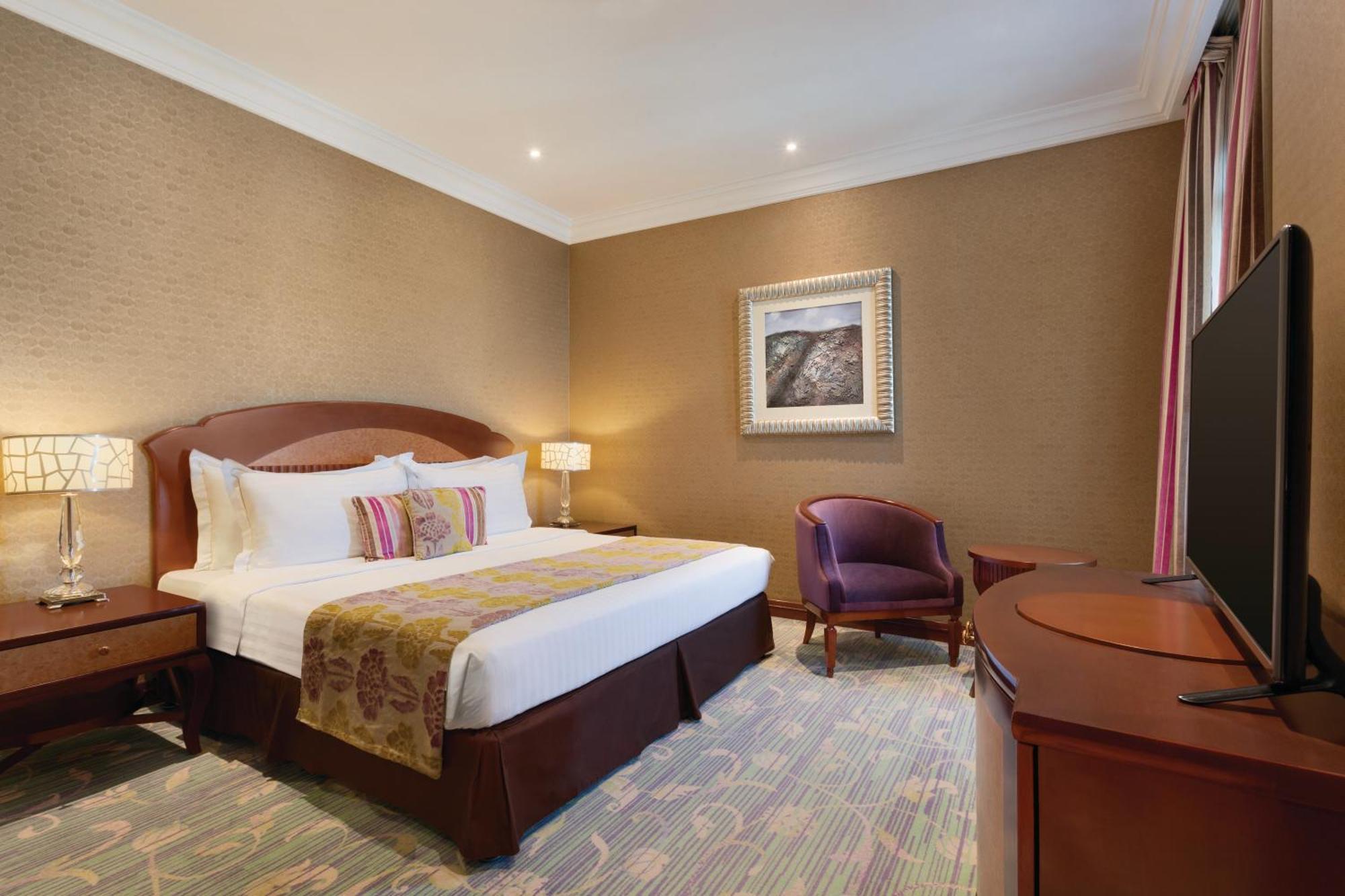 Grand Regency Doha, Trademark Collection By Wyndham Exterior photo The image shows a hotel room that features a cozy and elegant design. There's a large bed with white sheets and colorful decorative pillows. Next to the bed, there are nightstands with lamps. A comfortable armchair is positioned in the corner, and a 