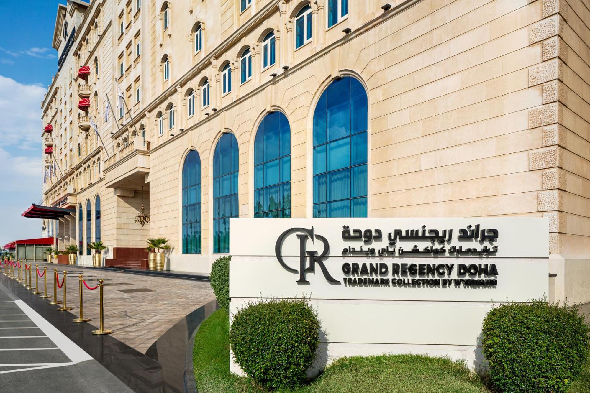 Grand Regency Doha, Trademark Collection By Wyndham Exterior photo The photo shows the entrance of the Grand Regency Doha, a luxury hotel. The building features a distinctive architectural style with large arched windows. There is a prominent sign featuring the hotel's name in both English and Arabic. The surroundin