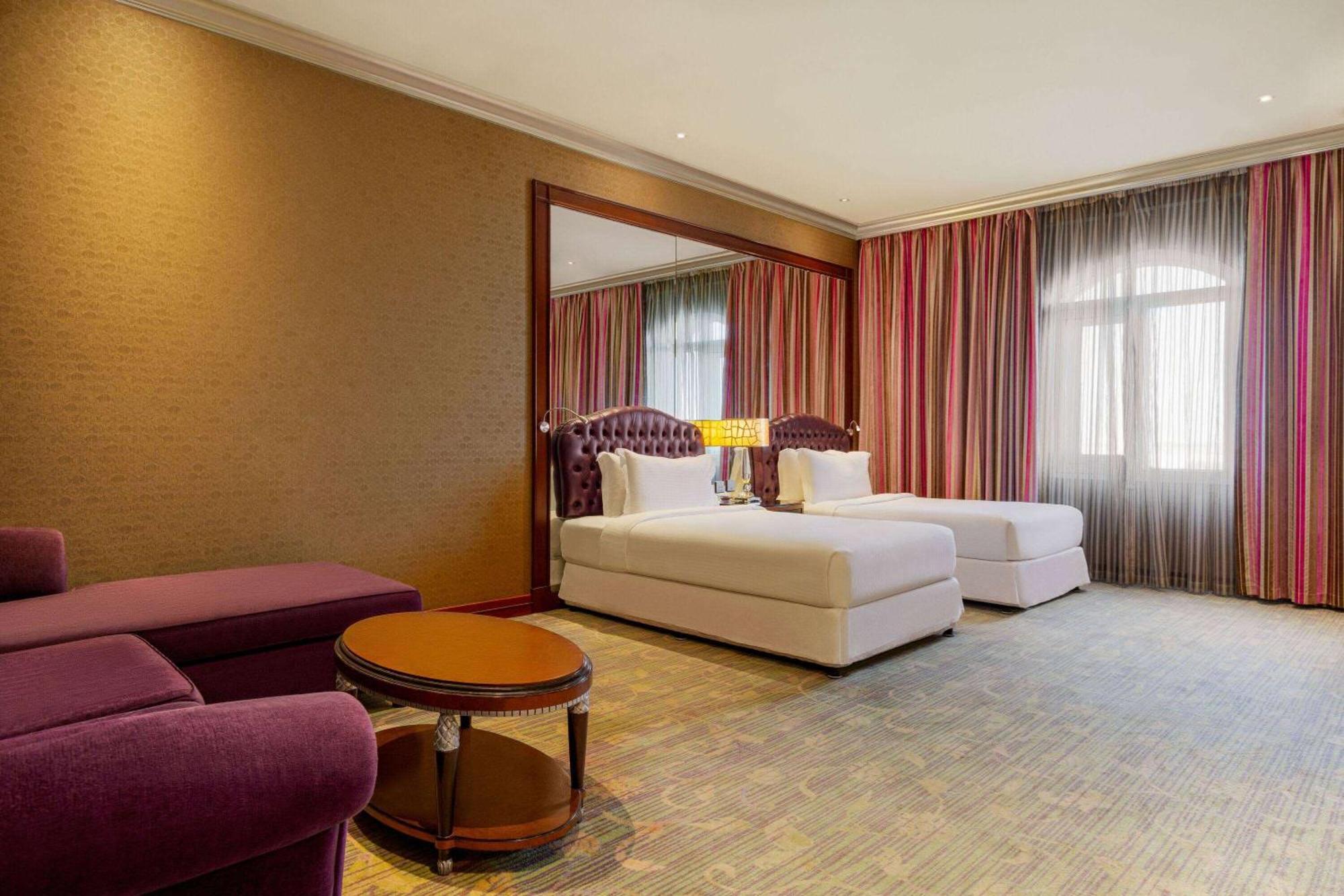 Grand Regency Doha, Trademark Collection By Wyndham Exterior photo The photo shows a hotel room featuring two beds with white bedding, a large mirror on one wall, and a window dressed with sheer and curtain panels. There is a small round table in the center of the room and a plush purple armchair nearby. The overall