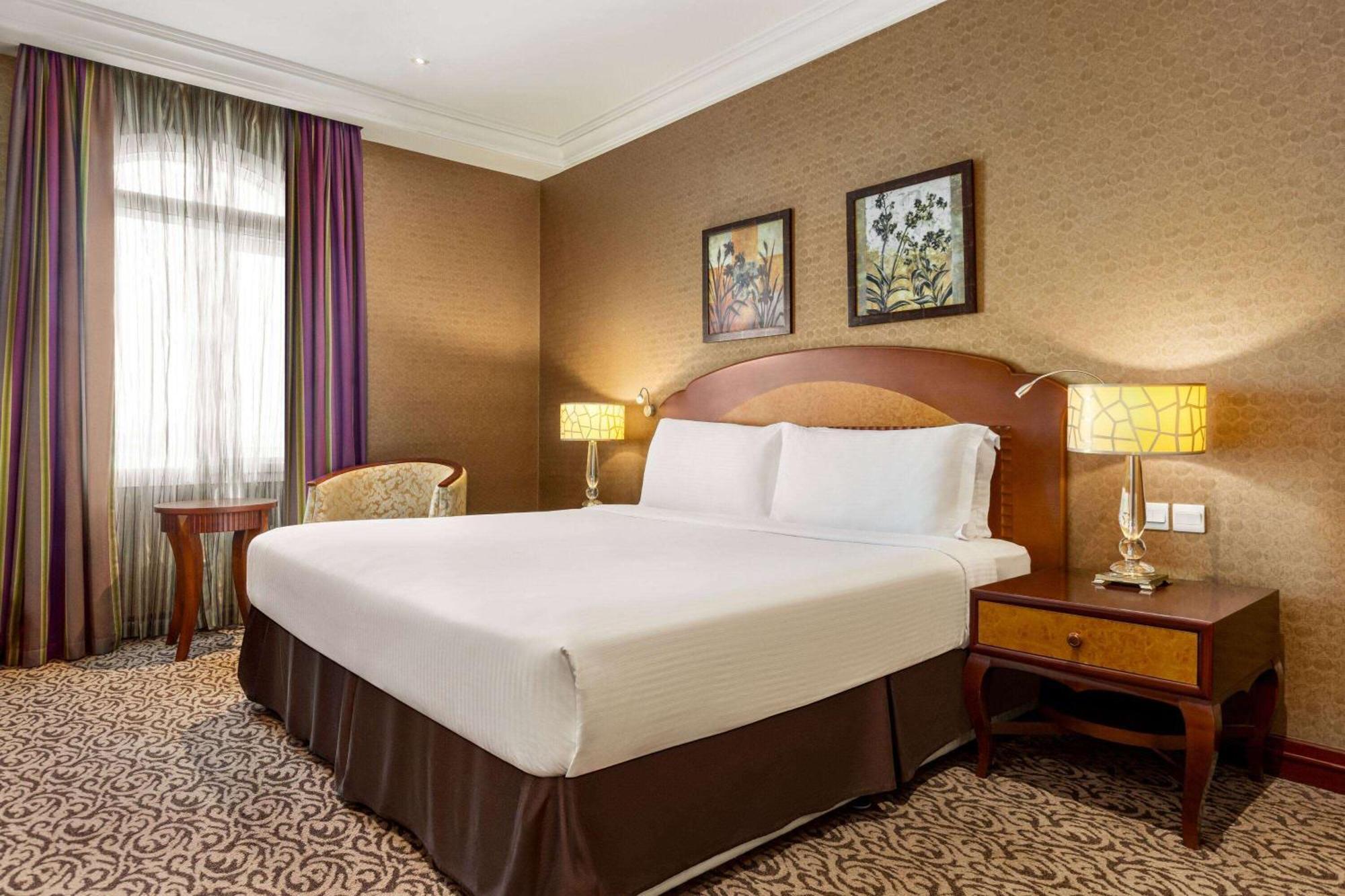 Grand Regency Doha, Trademark Collection By Wyndham Exterior photo The photo displays a comfortable hotel room featuring a large bed with white linens and a brown bed skirt. The room has a warm color palette, with textured brown walls and patterned carpet. There are two bedside lamps providing soft lighting next to 